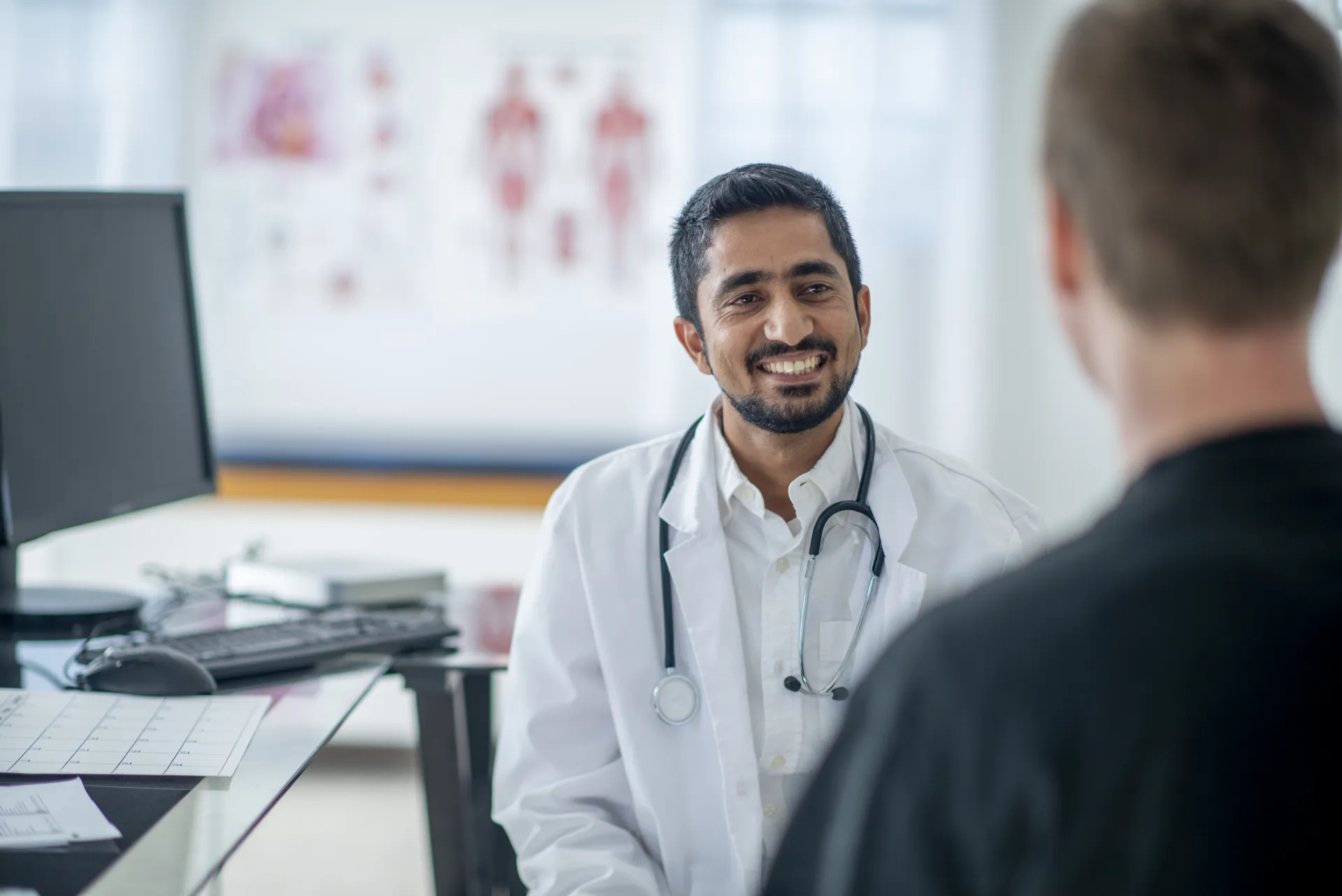 A doctor at his desk, computer beside him, smiling at the person across from him